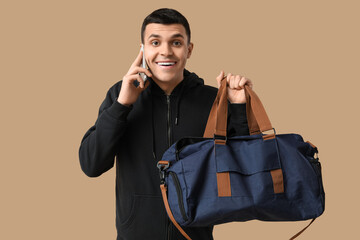 Young man with sports bag talking by phone on beige background