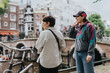 © qunica.com - Lesbian couple enjoying a casual day by a picturesque canal in Amsterdam, engaging in conversation and smiling. Represents love, joy, and adventure in an urban setting.