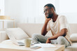 © SHOTPRIME STUDIO - Smiling African American man working on laptop in a modern living room The freelancer is sitting on a sofa, typing on his computer, surrounded by a cozy and stylish environment He appears happy and