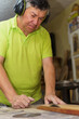 © fx-visual - A man in a green shirt is working on a project in a workshop. He is wearing earplugs and he is focused on his task cutting a wooden plank with a saw on the table