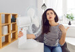 © Studio Romantic - Portrait of young upset brunette woman sitting at the desk at home and suffering from summer heat in front of fan. Female person using electric fan during heatwave in office on her workplace.