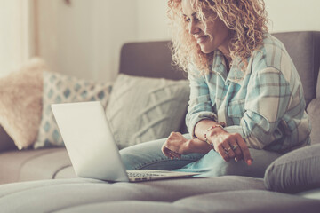 Happy blonde woman with curly hair using laptop sitting on sofa at home. Smiling 45 year old middle aged woman looking at computer while surfing internet, shopping on website and relaxing on living