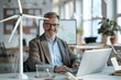 © Sourav Mittal - A smiling middle-aged business man sitting at his desk in a modern office using a laptop. A green wind turbine model sits next to him. The office has a light color scheme of white and light grey. The