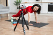 © sofiko14 - Woman in red workout attire doing push-ups on yoga mat, following online fitness tutorial at home. Exercise equipment like dumbbells and water bottle visible.