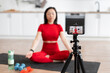 © sofiko14 - Woman meditating at home on yoga mat while recording video with smartphone. Front view of woman in red workout clothes filming meditation session. Focus on camera screen.
