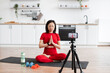 © sofiko14 - Woman in red sportswear meditating on yoga mat at home while recording session on camera. Modern kitchen background, fitness equipment on floor. Peaceful and focused atmosphere.