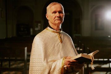 Sticker - Medium portrait of elderly Catholic priest wearing beige chasuble standing indoors with Bible in hands looking at camera