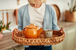 © Valerii Apetroaiei - Woman Holding Wicker Tray with Orange Teapot and Ceramic Mug in Cozy Kitchen Setting