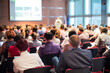 © kasto - Business and entrepreneurship symposium. Female speaker giving a talk at business meeting. Audience in conference hall. Rear view of unrecognized participant in audience.