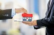 © Julio - Imagen de un experto en negocios inmoviliarios ajustándose la corbata con un modelo de casa sobre su escritorio.