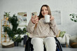 © Bliss - A young woman in a wheelchair sits at home, looking at her phone while holding a mug.