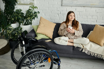 Wall Mural - A young woman in a wheelchair sits on a couch, holding a cup of coffee, while looking out the window.