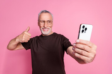 Wall Mural - Portrait of satisfied man with beard dressed t-shirt in glasses show thumb up make selfie on smartphone isolated on pink color background