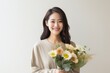 © Aksana - A young Asian woman smiling while arranging a bouquet of flowers on a table in a bright, modern room with large windows and potted plants, horizontal image. The flowers are a mix of vibrant colors and