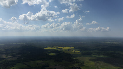 Poster -  A birds-eye perspective of a verdant landscape, dotted with green fields, beneath a clear blue sky adorned with fluffy white clouds, and punctuated by golden sunbe