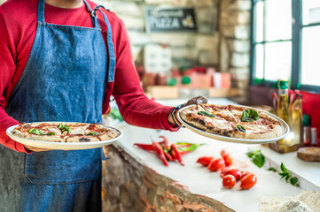 Chef holding two delicious pizzas in a rustic kitchen. Fresh ingredients and cozy ambiance highlight traditional Italian cuisine. Main focus on pizza at the left.