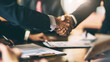 © Alena - A close-up photograph of a handshake sealing a business deal on a contract, with blurred office furniture and other hands holding pens in the background of a conference room.