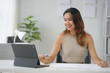 © amnaj - Smiling woman working on a tablet at her desk, engaged in remote work, surrounded by a modern office setup and greenery.