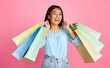© Prostock-studio - Asian young woman in a light blue blouse and jeans smiles brightly as she holds up a stack of colorful shopping bags against a pink backdrop