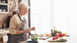 © Prostock-studio - A woman wearing an apron stands in a modern kitchen, preparing a fresh salad. The kitchen has a brick backsplash, white cabinets, and a large window that lets in natural light, copy space