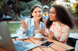 © Prostock-studio - Two women are sitting at a table at an outdoor cafe, using a laptop and phone. They are both smiling and looking at each other. There is a laptop and two phones on the table.