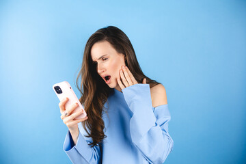 Wall Mural - Mad and shock young brunette beautiful woman using mobile phone isolated on blue background. Yelling unhappy woman hold mobile phone. Disappointed sad upset lady horrified impressed news. WTF. Oh no.