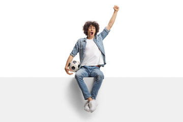 Happy young man cheering and holding a football seated on a blank panel