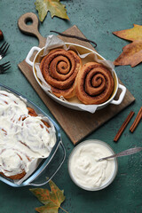 Wall Mural - Baking dishes of tasty cinnamon rolls with cream and autumn leaves on green background