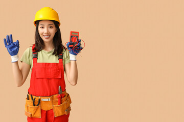 Wall Mural - young female electrician with multimeter showing ok gesture on beige background