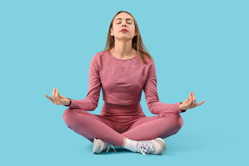 Wall Mural - Beautiful young woman in sportswear meditating on blue background