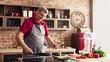 © Prostock-studio - A smiling senior man wearing an apron cooks in a modern kitchen with brick walls. He is stirring food in a frying pan on the stovetop with a wooden spoon.