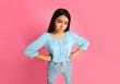 © Prostock-studio - Asian young woman standing with her hands on her hips, looking disappointed. She is wearing a light blue blouse and light blue jeans. The background is a solid pink color.