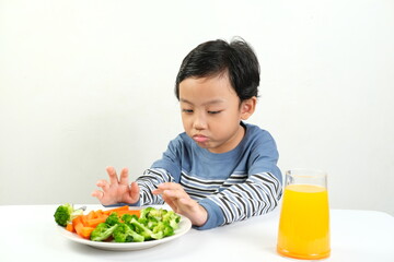A young boy refuses a plate of vegetables and a glass of orange juice, showing displeasure. This captures the common struggle of getting kids to eat healthy food.