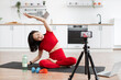 © sofiko14 - Woman practicing yoga in kitchen, stretching on fitness mat. Recording video with smartphone on tripod. Home workout with water bottle, dumbbells, resistance bands nearby.