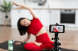 © sofiko14 - Woman in red activewear filming yoga session at home with smartphone. Focus on phone screen displaying stretching exercise. Home workout, fitness routine, and online yoga class.
