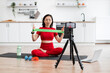 © sofiko14 - Woman in red workout attire demonstrating resistance band exercise for online fitness class in home kitchen, seated on yoga mat with water bottle, dumbbells, smartphone tripod, and laptop.