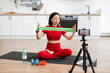 © sofiko14 - Woman in red workout clothes demonstrating resistance band exercises during an online fitness class in modern kitchen. Filming fitness video with smartphone and tripod setup.
