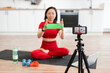 © sofiko14 - Woman in red workout clothes filming online fitness class at home using resistance band. Fitness instructor demonstrating exercises for virtual audience. Home workout setup with tripod, smartphone.