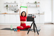 © sofiko14 - Woman in red activewear using resistance band for exercise during online fitness class in home kitchen setting. Yoga mat, water bottle, and dumbbells visible, indicating healthy lifestyle.