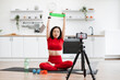 © sofiko14 - Woman in red athletic wear using resistance band for home workout. Sitting on yoga mat in modern kitchen, filming exercise routine. Fitness equipment and water bottle nearby.