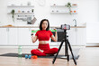 © sofiko14 - Woman in red sportswear demonstrating fitness exercises on camera in modern kitchen setting. Green resistance band, water bottle, yoga mat, and laptop create home workout environment.