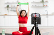 © sofiko14 - Woman in red workout attire stretching with resistance band while filming online workout video. Exercise takes place in a modern home setting with camera on tripod focusing on her.