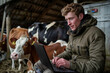 © Peeradontax - A young farmer in a barn, wearing a warm jacket, works on a laptop while a curious brown and white cow looks on.