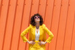 © VISTA by Westend61 - Woman with arms akimbo wearing sunglasses and yellow blazer in front of orange wall on sunny day