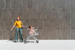 © VISTA by Westend61 - Happy mother with daughter sitting inside shopping cart near wall