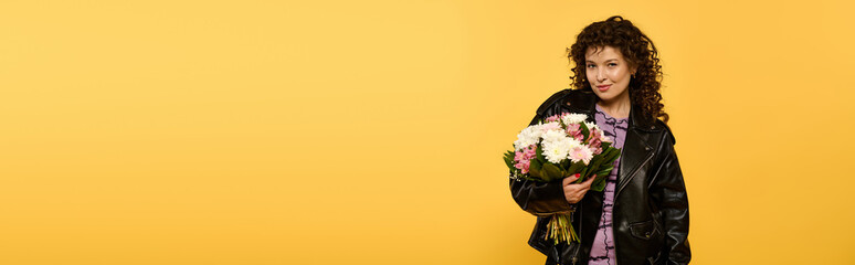 Wall Mural - A stylish young woman in a black leather jacket poses against a yellow background while holding a bouquet of flowers.