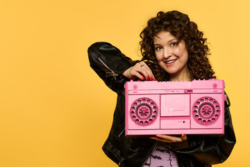 Wall Mural - Woman with curly hair smiles, holding pink boombox against yellow background, adorned in black leather jacket.