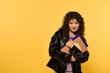 © LIGHTFIELD STUDIOS - A stylish young woman with curly hair, wearing a black leather jacket, poses against a yellow background while holding a stack of books.