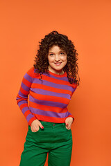 Wall Mural - A young woman with curly hair poses against a bright orange background, wearing a red and purple striped shirt and green pants.