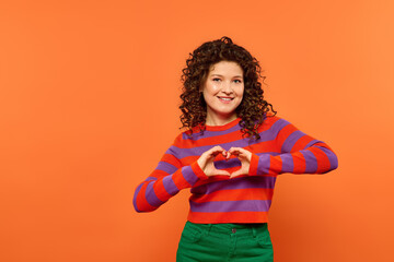 Wall Mural - A young woman with curly hair poses against a bright orange background, forming a heart shape with her hands.